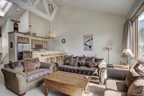 A cozy living room features two sofas, a coffee table, a kitchen area with wooden countertops, and a bar counter. The room is decorated with a bear sculpture and posters, with large windows allowing natural light.