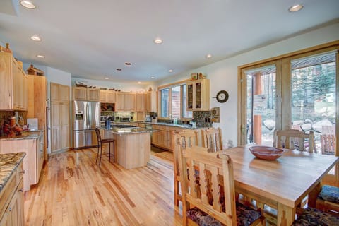 A spacious kitchen with light wood cabinets, stainless steel appliances, granite countertops, wood flooring, and an adjacent wooden dining table set near large glass doors.