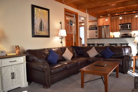 A cozy living room with a brown leather sectional sofa, decorated with pillows. There is a framed painting on the wall, a wooden coffee table, and a view of the kitchen in the background.