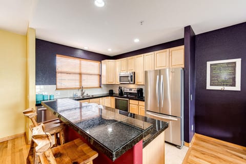 Modern kitchen with wooden cabinets, stainless steel appliances, granite countertops, and a central island with bar stools. Walls are painted in yellow and dark purple tones.