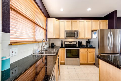 A modern kitchen with light wood cabinets, stainless steel appliances, a double sink, granite countertops, tiled floor, and large window with wooden blinds.
