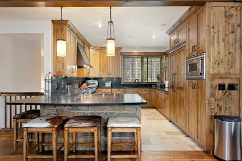 A rustic kitchen features wood cabinets, stainless steel appliances, an island with seating, and stone countertops. Natural light enters through a window by the sink.