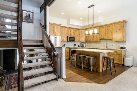 Modern kitchen with wooden cabinets, an island with stools, and stainless steel appliances. To the left, a staircase leads to an upper floor, and a framed portrait hangs on the wall above the steps.