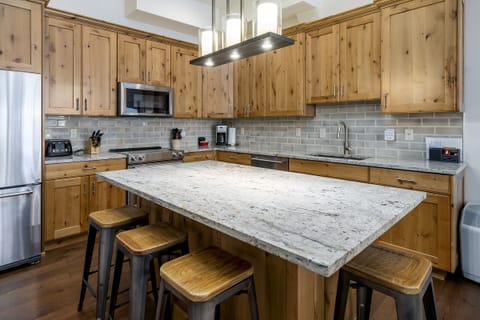 Spacious modern kitchen with light wood cabinets, stainless steel appliances, subway tile backsplash, and large granite island with four wooden stools.