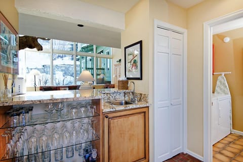 A home bar area with granite countertops features a sink, glass shelving filled with various glassware, and light wood cabinetry, adjacent to a hallway leading to a laundry room with a washer and dryer.