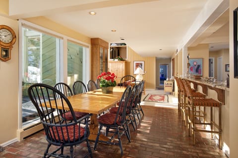 A dining room with a wooden table, black spindle-back chairs, and a potted plant centerpiece. Brick flooring extends to a connected kitchen with a counter and bar stools. Large windows provide natural light.