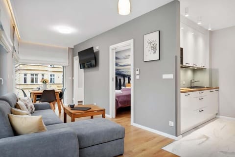 A bright living room featuring a comfortable gray sofa, a wooden coffee table, and a modern, open-plan layout with the kitchen visible in the background.