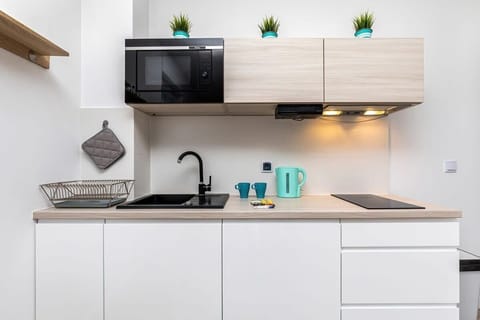 A clean and minimalist kitchen with white cabinets, wooden shelving, and teal accents. The setup includes basic kitchen appliances and plenty of storage space.