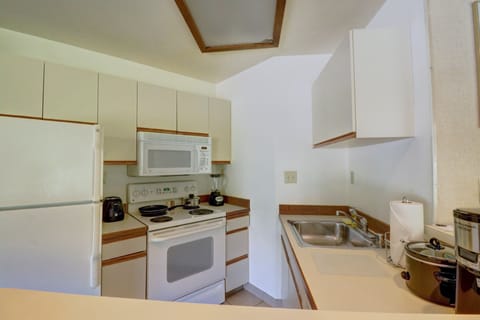 A compact kitchen with white cabinets, a white refrigerator, microwave, and electric stove. The countertop holds a coffee maker, toaster, paper towel roll, and various small appliances.