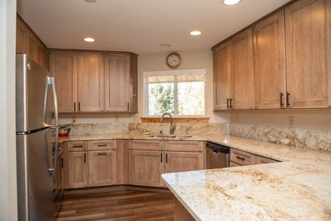 A modern kitchen with wooden cabinets, stainless steel refrigerator and dishwasher, granite countertops, and a window above the sink.