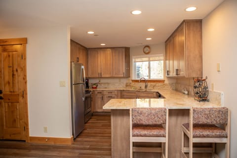 A modern kitchen with wooden cabinets, granite countertops, stainless steel appliances, a small window above the sink, and a breakfast bar with two padded stools.