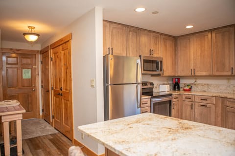 Modern kitchen with wooden cabinets, stainless steel appliances including a fridge and microwave, granite countertops, and a kitchen island. Doorway and hallway visible in the background.