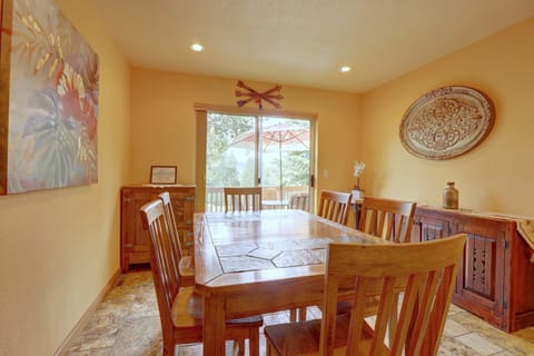 A wooden dining table with six chairs sits in a brightly lit room with an ornate wall decoration and a large sliding glass door leading to an outdoor area. Art and furniture adorn the walls and floors.