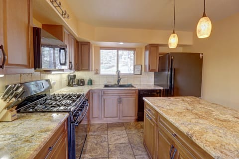 A modern kitchen with wooden cabinets, granite countertops, stainless steel appliances, hanging pendant lights, and a center island. A window above the sink provides natural light.