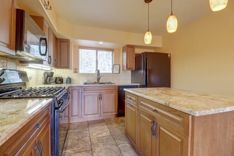 A modern kitchen features wooden cabinets, granite countertops, a gas stove, stainless steel appliances, and pendant lights above the island. The floor is tiled, and a window provides natural light.