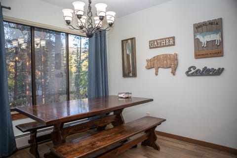 A dining area with a wooden table and benches, wall art featuring a cow, a pig, and the words "Gather" and "Eatery." A large window with blue curtains overlooks trees. A chandelier hangs above the table.