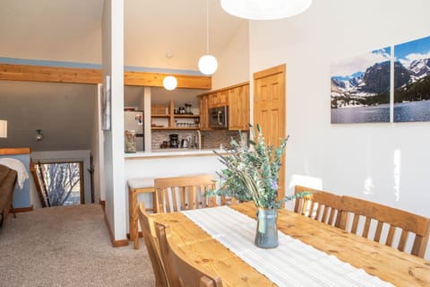 A cozy dining area with a wooden table and chairs, adjacent to a small kitchen with light wood cabinetry. The space is decorated with nature-themed artwork and lit by round pendant lights.