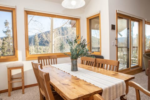A wooden dining table with a decorative vase of flowers is placed near large windows offering a scenic view of a mountainous landscape. The room features light-colored walls and wooden furniture.