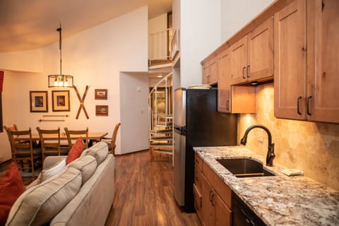 Interior view of a cozy kitchen and living area with wooden cabinets, granite countertops, and an adjacent dining table. A spiral staircase and framed artwork are visible in the background.