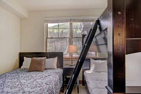 A bedroom featuring a double bed and a bunk bed with a ladder. The room has a large window with shades, offering a view of a forested hillside. A bedside table with a lamp is placed between the beds.