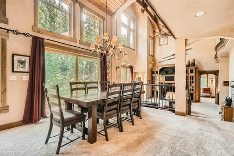 Spacious dining area with a large wooden table, high vaulted ceilings, chandelier, large windows, and carpeted floors, leading to a living room with a balcony and built-in shelves.