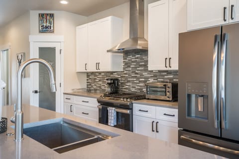 Modern kitchen with white cabinetry, stainless steel appliances, including a double-door fridge and oven, and a gray tiled backsplash. The countertop features a double sink with a high-arc faucet.