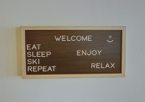 A wooden-framed sign with white letters reads, "WELCOME. EAT SLEEP SKI REPEAT ENJOY RELAX" on a brown background. There is a smiley face next to the word "WELCOME.