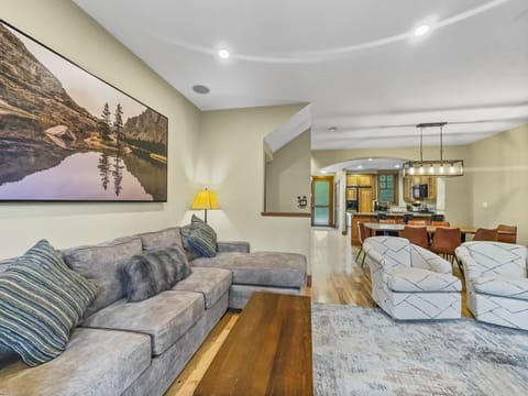 A living room with a gray sectional sofa, patterned chairs, wooden coffee table, wall art of a lake scene, and a view into a kitchen and dining area with a light fixture and brown dining chairs.