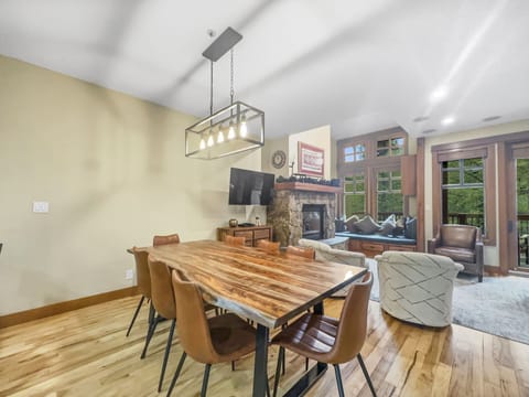 A modern dining room with a wooden table, six brown chairs, a large rectangular light fixture, and an adjacent living area with a fireplace, cozy seating, and large windows.