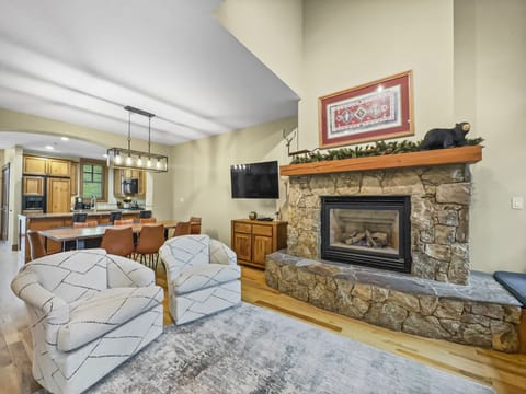 A cozy living room with a stone fireplace, two striped armchairs, and a mounted TV. The adjacent kitchen features wooden cabinets and a countertop with four bar stools. The space is adorned with warm tones.