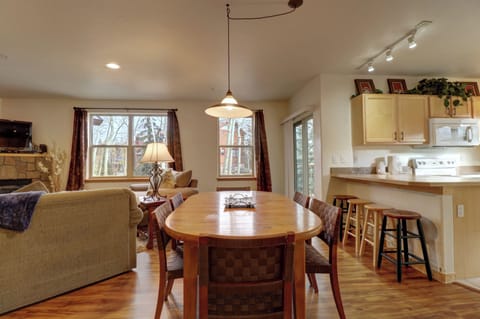 A cozy living and dining area with a wooden table, four chairs, a sofa, and a TV next to a fireplace. The open kitchen has light wood cabinets, a microwave, a stove, and bar stools at the counter.