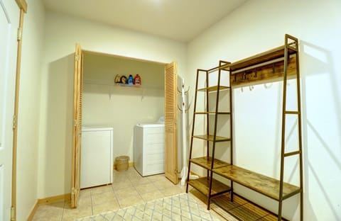 A laundry room with a washer, dryer, shelf with cleaning supplies, a wooden coat rack, and a metal shelving unit. The tiled floor has a patterned rug. The room is well-lit with white walls.