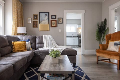 Spacious living room with a plush gray sectional adorned with vibrant accent pillows next to a geometric rug and rustic coffee table. Sunlight streams through large windows, highlighting accent chairs and gallery wall art. 🛋️🌟🖼️📺