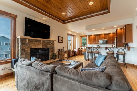Living area with large mounted flat screen smart TV, gas fireplace with beautiful stone surround, hardwood flooring and ample seating.