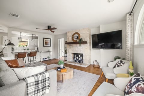 A large TV is mounted to the side of the fireplace, blending functionality with style. Two white, mid-century modern armchairs sit in front of a large, arched window. 