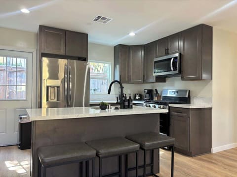 Modern kitchen with sleek dark cabinetry, stainless steel appliances, and a spacious quartz countertop with high top seating. Features a gas stove, large fridge, microwave, and black pull-down faucet. Bright, inviting, and perfect for group meals.