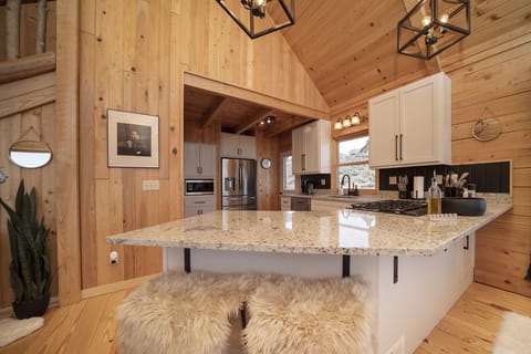 A modern kitchen with wooden walls and ceiling, a granite island with cushioned stools, stainless steel appliances, and white cabinets