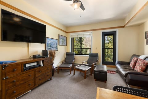 Living room with a mounted flat-screen TV, two lounge chairs with footrests, a black leather sofa, a wooden chest of drawers, and a balcony with a view of trees.