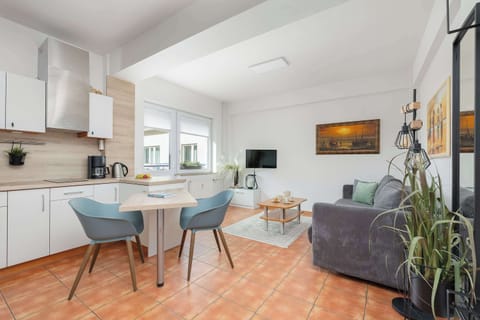 A modern kitchen with white cabinets, a gray couch in the living area, and a dining table with blue chairs. A plant is visible in the foreground.