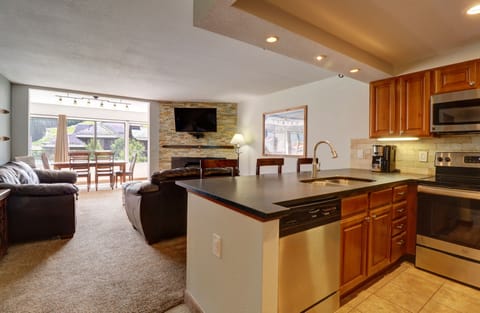 Open-concept kitchen and living area with stainless steel appliances, wooden cabinets, and a stone accent wall. The living area features a wall-mounted TV and leather seating with views of a patio outside.