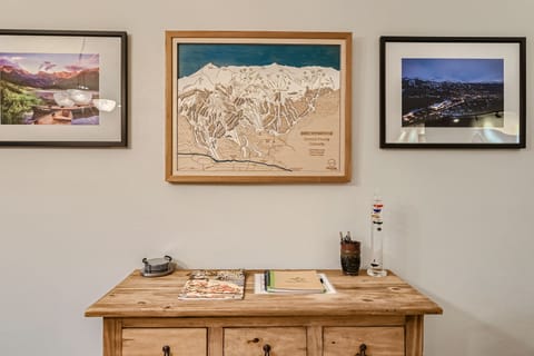 A wooden cabinet with a few magazines and small objects sits below a framed map of Breckenridge and two framed photographs of mountainous landscapes on a beige wall.
