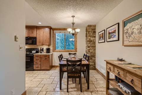 A small dining area with a wooden table and four chairs, adjacent to a kitchen. The space has stone tile flooring, a window, and decorated walls. A chandelier hangs above the table.