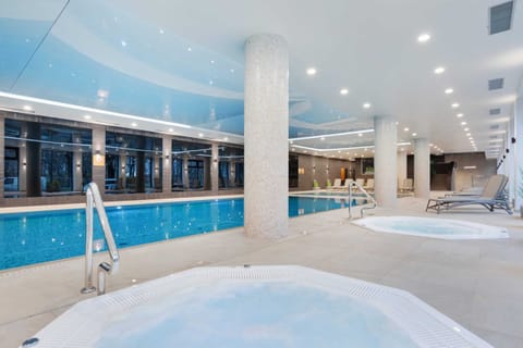 A luxurious indoor swimming pool area with a jacuzzi, surrounded by lounge chairs.
