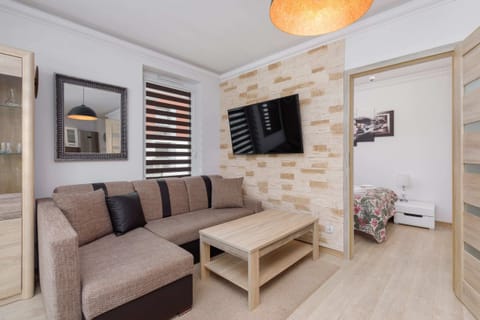 A cozy living room featuring a grey sofa with a wooden coffee table, a flat-screen TV mounted on the wall, and decorative wall panels with a stone-like design.