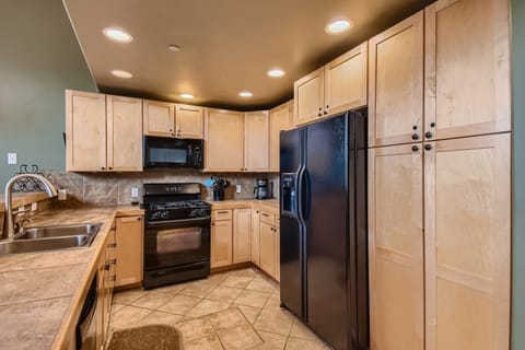 A modern kitchen with light wood cabinets, black appliances, including a refrigerator and oven, tile countertops, and recessed lighting.