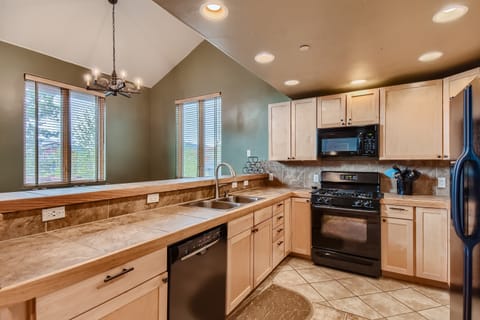 A modern kitchen with wooden cabinets, black appliances, a double sink, and tiled countertops. There are large windows with blinds, a chandelier, and tile flooring.