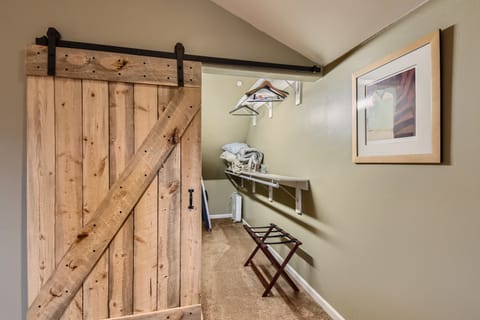 A small walk-in closet with beige walls, a wooden sliding barn door, a luggage rack, hanging clothes, a shelf, and a framed picture on the right wall.