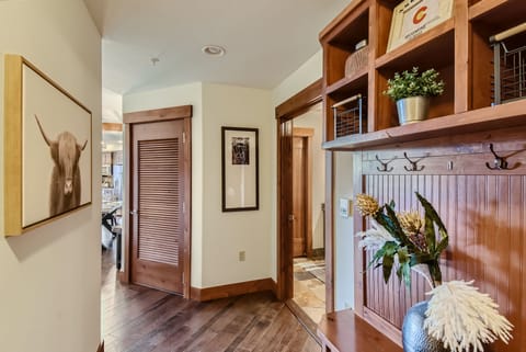 A hallway features wooden accents, a painting of a cow, potted plant, coat hooks, and storage shelves. A louvered door opens to the left, and a glimpse of another room is visible through an open doorway.