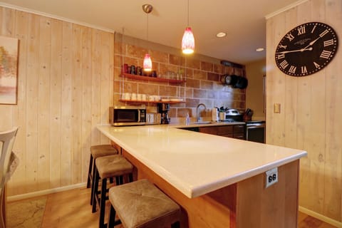 A cozy kitchen with wooden walls, a large wall clock, and a white countertop with three bar stools. Pendant lights hang above the counter, and kitchen appliances are visible in the background.
