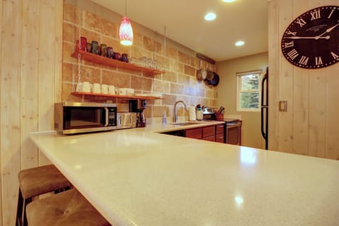 A modern kitchen with a long countertop, microwave, sink, and various mugs on shelves. A large clock is mounted on the wooden wall, and a window lets in natural light.